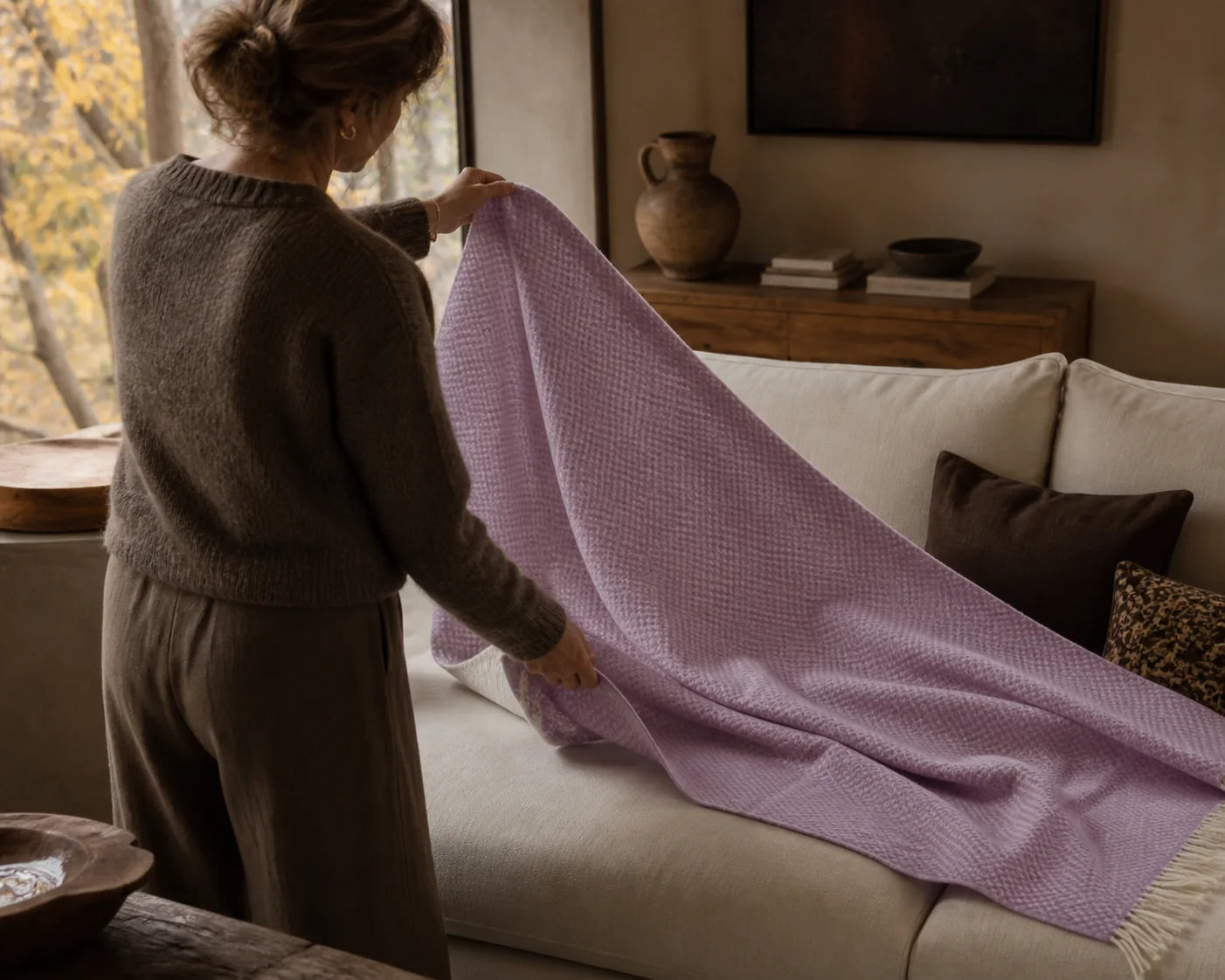 A woman aged 30 to 50 seen from behind opening a mauve Sofia Toscano throw across a living-room sofa