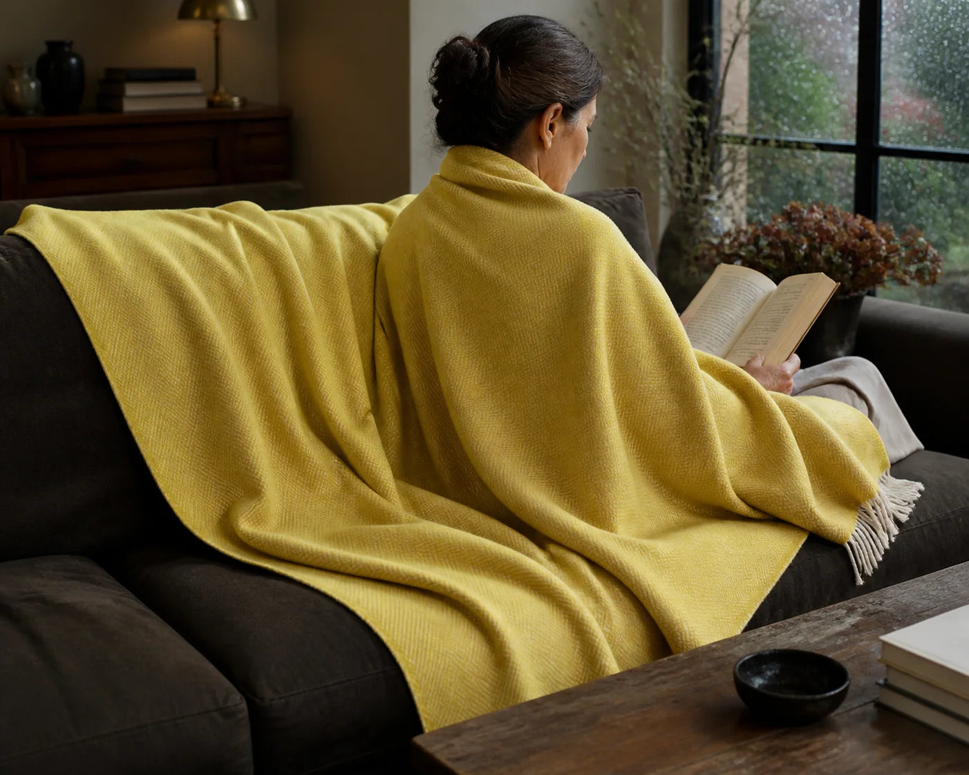 A woman aged 30 to 50 seen from behind reading with an open mustard Sofia Toscano throw in her living room